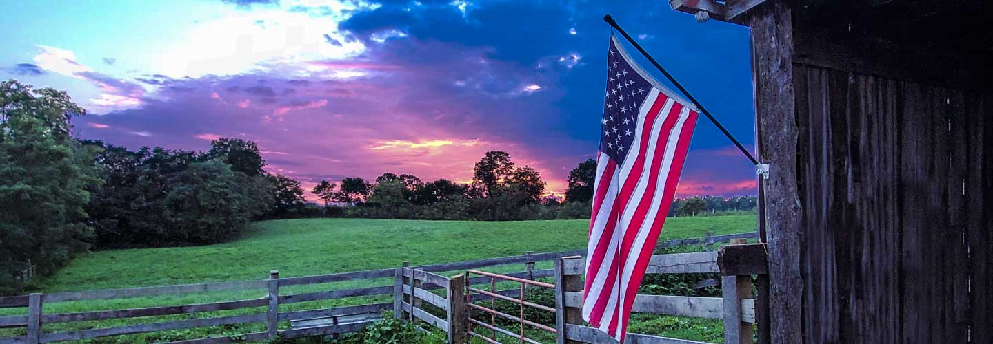 An American flags hangs from the side of a rustic barn with a beautiful sunset in the background